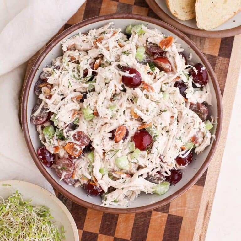 A bowl of homemade chicken salad with red grapes and fresh dill on a checkered cutting board next to toast and sprouts.