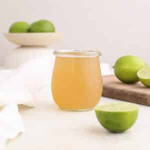 A jar of homemade lime syrup on a cutting board next to fresh limes.