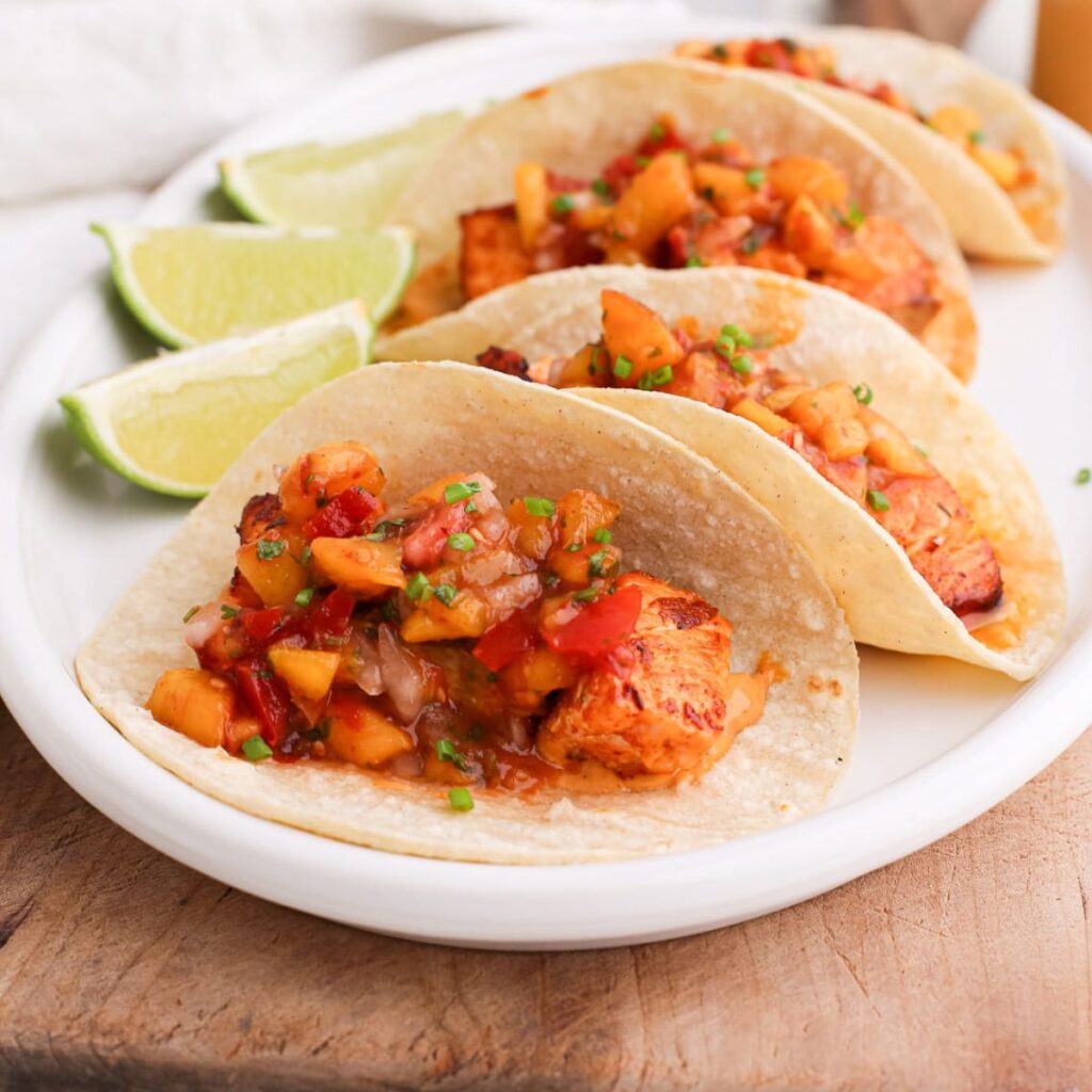 Blackened salmon tacos topped with peach salsa next to lime wedges.