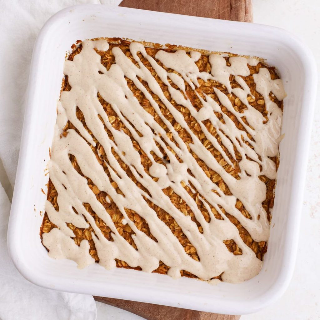 A square ceramic pan of pumpkin baked oatmeal with a maple yogurt glaze drizzled on top next to a bowl of pumpkin puree.
