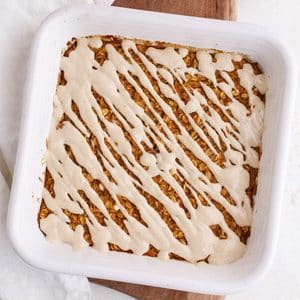 A square ceramic pan of pumpkin baked oatmeal with a maple yogurt glaze drizzled on top next to a bowl of pumpkin puree.
