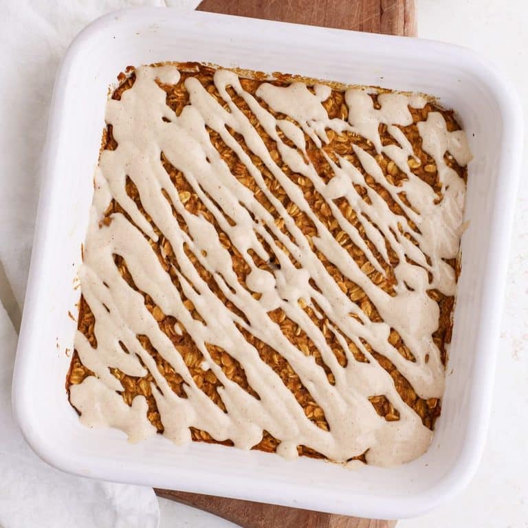 A square ceramic pan of pumpkin baked oatmeal with a maple yogurt glaze drizzled on top next to a bowl of pumpkin puree.